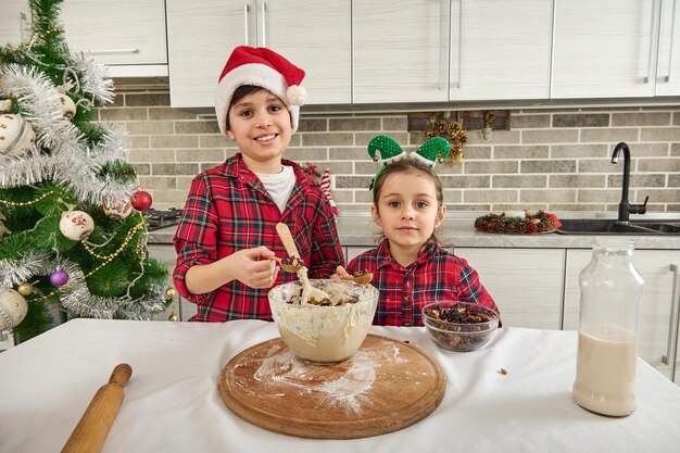 Lindas crianças europeias, lindo menino pré-adolescente alegre e sua irmã mais nova, linda menina olhando para a câmera em pé ao lado da mesa da cozinha e cozinhando a massa de bolos de Natal