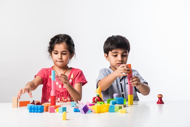 Lindas crianças indianas asiáticas brincando com brinquedos de plástico coloridos ou blocos enquanto estão sentadas à mesa ou isoladas sobre um fundo branco