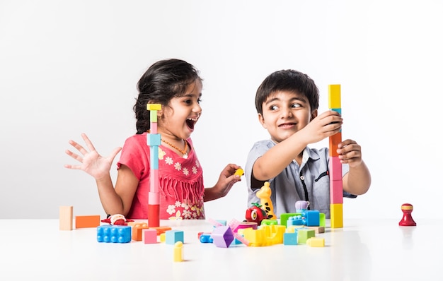 Lindas crianças indianas asiáticas brincando com brinquedos de plástico coloridos ou blocos enquanto estão sentadas à mesa ou isoladas sobre um fundo branco