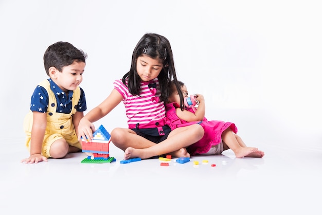 Lindas crianças indianas ou asiáticas brincando com brinquedos ou blocos e se divertindo enquanto estão sentadas à mesa ou isoladas sobre um fundo branco