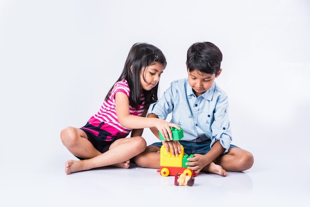 Lindas crianças indianas ou asiáticas brincando com brinquedos ou blocos e se divertindo enquanto estão sentadas à mesa ou isoladas sobre um fundo branco