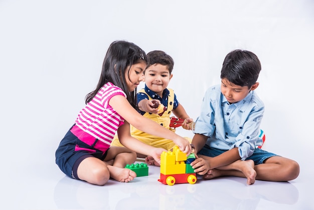 Lindas crianças indianas ou asiáticas brincando com brinquedos ou blocos e se divertindo enquanto estão sentadas à mesa ou isoladas sobre um fundo branco