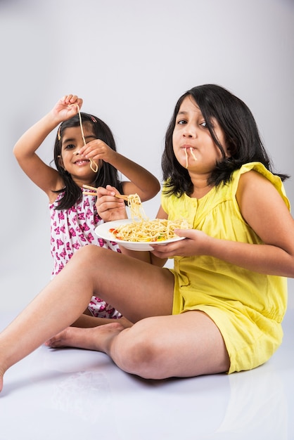 Lindas garotas indianas ou asiáticas brincalhonas comendo macarrão chinês saboroso com garfo ou pauzinhos, isolado sobre um fundo colorido