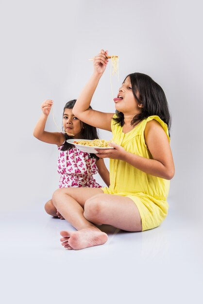 Lindas garotas indianas ou asiáticas brincalhonas comendo macarrão chinês saboroso com garfo ou pauzinhos, isolado sobre um fundo colorido