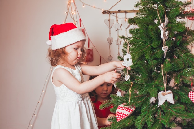 Lindas hermanas con gorros de Santa Claus están decorando el árbol de Navidad
