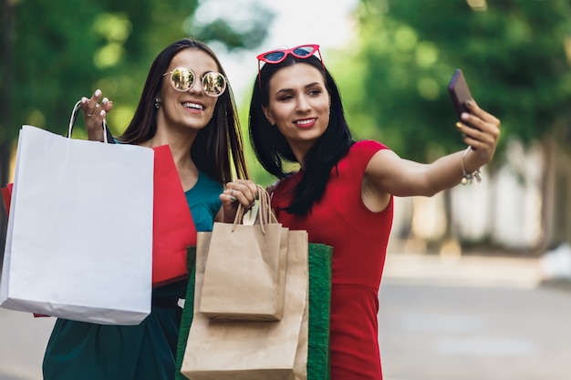 Lindas meninas felizes em óculos de sol segurando sacolas de compras, fazendo selfie telefone inteligente e sorrindo