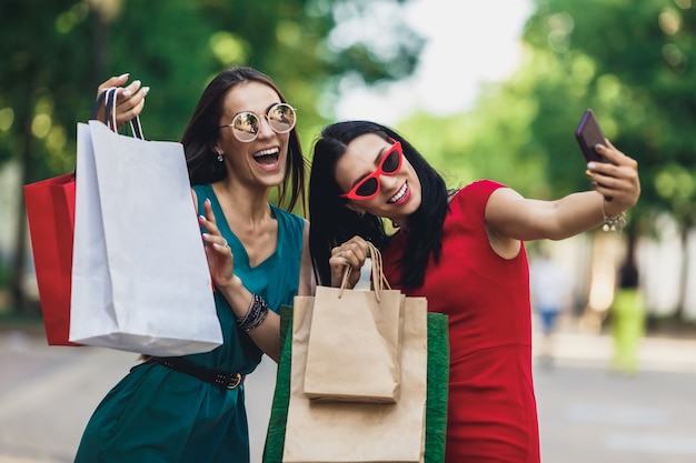 Lindas meninas felizes em óculos de sol, segurando sacolas de compras, fazendo selfie telefone inteligente e sorrindo.