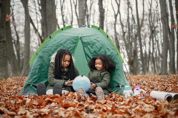 Lindas meninas negras na barraca acampando na floresta Duas irmãzinhas sentadas em uma barraca na floresta de outono e olhando para um globo Meninas negras vestindo casacos cáqui