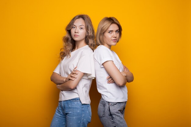 Lindas mujeres rubias posando espalda con espalda en una pared amarilla con las manos cruzadas