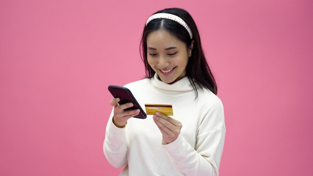 Lindas mulheres asiáticas sorriem usando smartphone e cartão de crédito comprando compras on-line e animadas com pagamento bem-sucedido em fundo rosa isolado