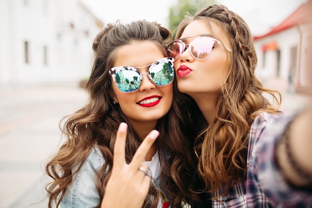 Lindas namoradas morenas com penteado, óculos de sol espelhados e lábios vermelhos fazendo selfie com cara de pato.