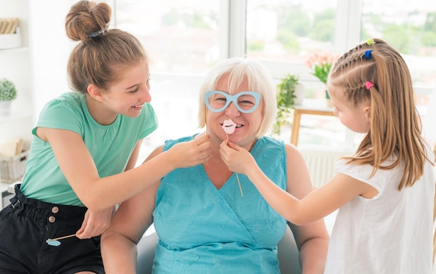 Lindas nietas poniendo vasos de papel y labios en abuela sonriente en casa