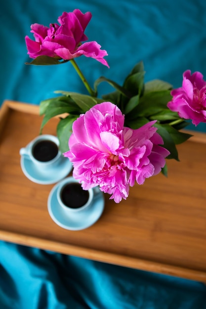 Lindas peônias rosa em um vaso de vidro e duas xícaras de café estão em uma bandeja de madeira na cama