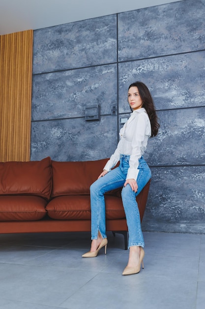 Lindas pernas de mulher esbelta em sapatos clássicos bege com saltos finos e jeans Garota elegante de salto senta-se em um sofá de couro marrom