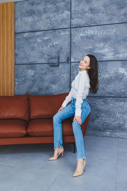 Lindas pernas de mulher esbelta em sapatos clássicos bege com saltos finos e jeans Garota elegante de salto senta-se em um sofá de couro marrom