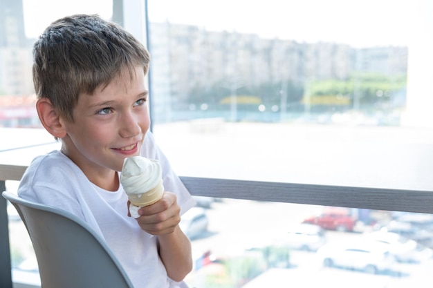 Un lindo adolescente come un helado blanco arremolinado en una taza de gofres sentado en una mesa junto a la ventana en un café