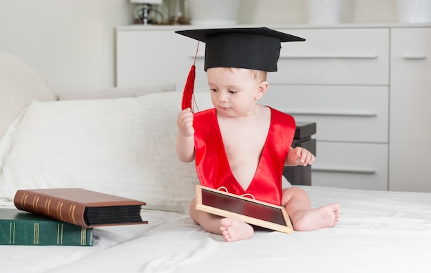 Lindo bebé de 10 meses en gorro de graduación sosteniendo tableta digital y mirando la pila de libros