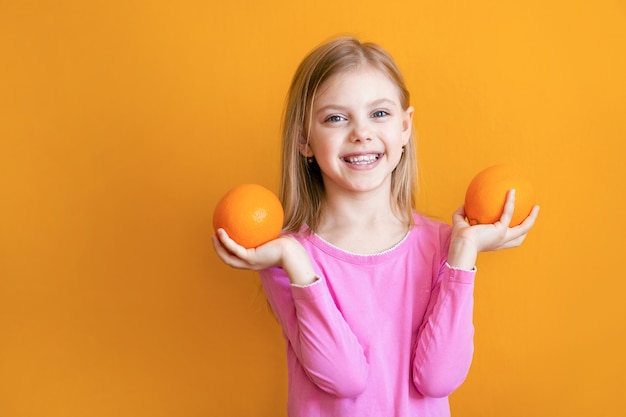 Lindo bebé de 8 años en una pared de color naranja, una niña bonita rubia con appelsins en sus manos sonríe