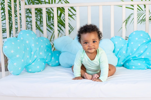 Lindo bebé africano sentado en la cama en casa antes de acostarse en la cama azul, el concepto de sueño saludable y accesorios de algodón para ropa de cama de bebé.