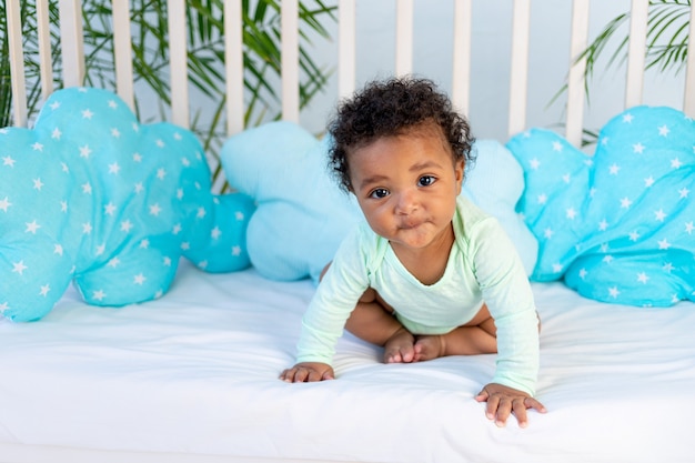 Lindo bebê africano sentado na cama em casa antes de ir para a cama na cama azul, o conceito de sono saudável e acessórios de algodão da cama de bebê.