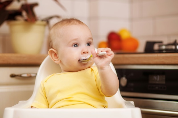 Lindo bebé aprende a comer con una cuchara sentado en una silla en la cocina.