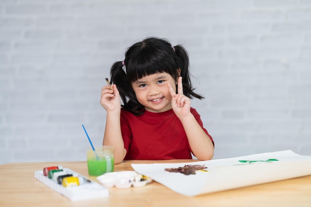 Lindo bebé asiático sonriendo y mostrando dos dedos de la mano y pintando con pinturas de colores usando acuarela Chica asiática usando pincel de dibujo color Concepto de estilo de vida de actividad de bebé