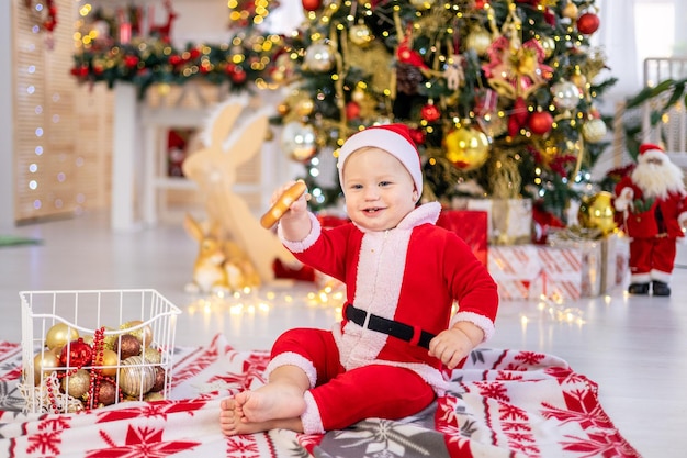 Un lindo bebé disfrazado de Papá Noel está sentado con juguetes de árbol de Navidad bajo un árbol de Navidad festivo de la casa un niño feliz está celebrando la Navidad y el Año Nuevo en casa