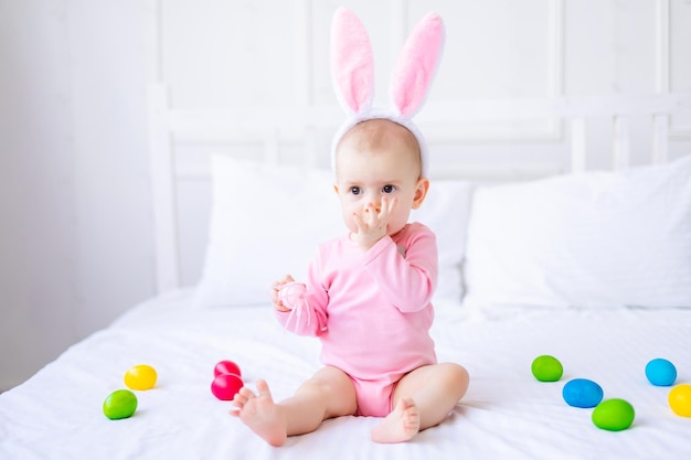 Lindo bebé divertido con orejas de conejo y coloridos huevos de Pascua en casa en una cama de ropa de cama blanca Tarjeta de felicitación de Pascua