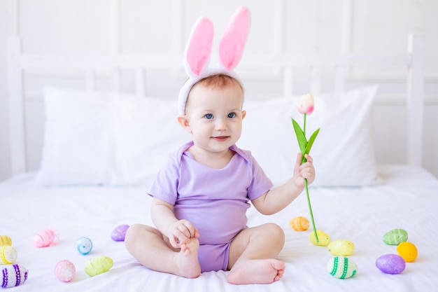 Lindo bebé divertido con orejas de conejo con un tulipán en la mano y coloridos huevos de Pascua en casa en una cama de ropa de cama blanca Tarjeta de felicitación de Pascua