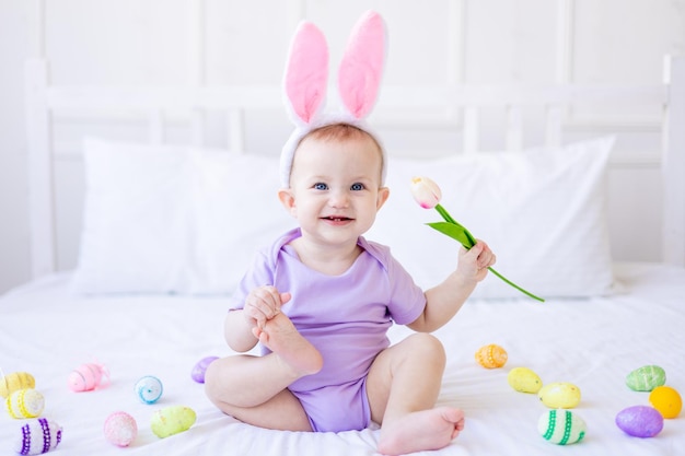 Lindo bebé divertido con orejas de conejo con un tulipán en la mano y coloridos huevos de Pascua en casa en una cama de ropa de cama blanca Tarjeta de felicitación de Pascua