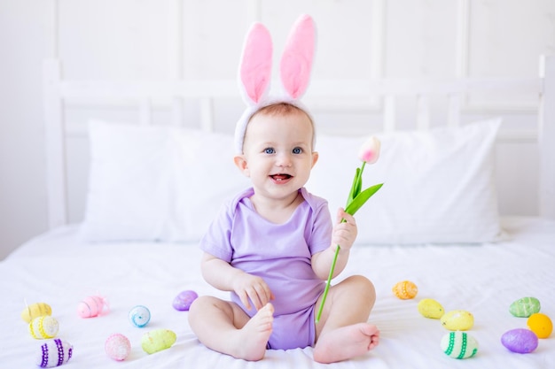 Foto lindo bebé divertido con orejas de conejo con un tulipán en la mano y coloridos huevos de pascua en casa en una cama de ropa de cama blanca tarjeta de felicitación de pascua