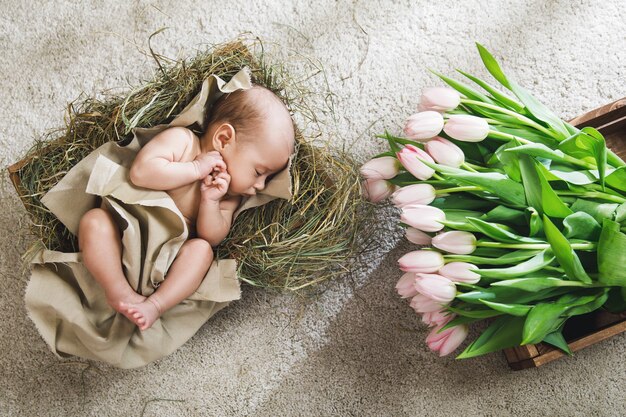 Lindo bebé está acostado en la caja de madera llena de heno y montón de tulipanes rosas
