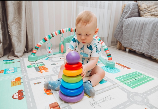 Un lindo bebé está jugando con una pirámide de juguete del arco iris mientras está sentado en una alfombra de juego en un dormitorio soleado Juguetes para niños pequeños
