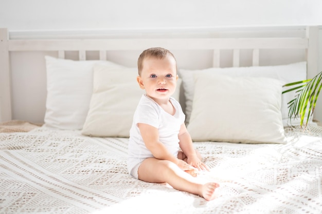 Un lindo bebé está sentado en una cama con sábanas blancas en el dormitorio de la casa. El retrato de un niño mira a la cámara.