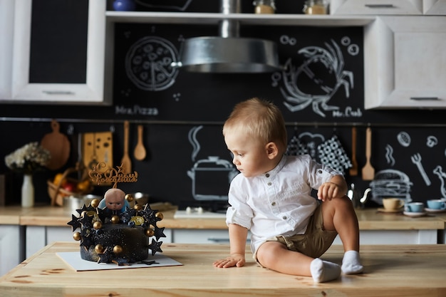 Lindo bebé con estilo en camisa blanca y pantalones cortos se sienta en la mesa, mirando un pastel de cumpleaños y posando en el interior de la cocina