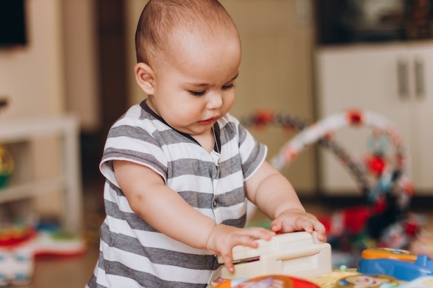 Lindo bebê gordinho fica de pé e brinca com uma mesa de música infantil. muitos brinquedos coloridos em todos os lugares.