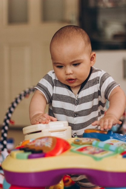 Foto lindo bebê gordinho fica de pé e brinca com uma mesa de música infantil. muitos brinquedos coloridos em todos os lugares.