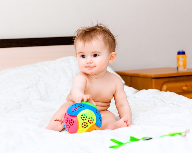 Lindo bebê loiro brincando com uma bola, sentado na cama no quarto. bebê feliz de 6 meses brincando com uma bola
