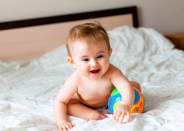 Lindo bebê loiro brincando com uma bola, sentado na cama no quarto. bebê feliz de 6 meses brincando com uma bola