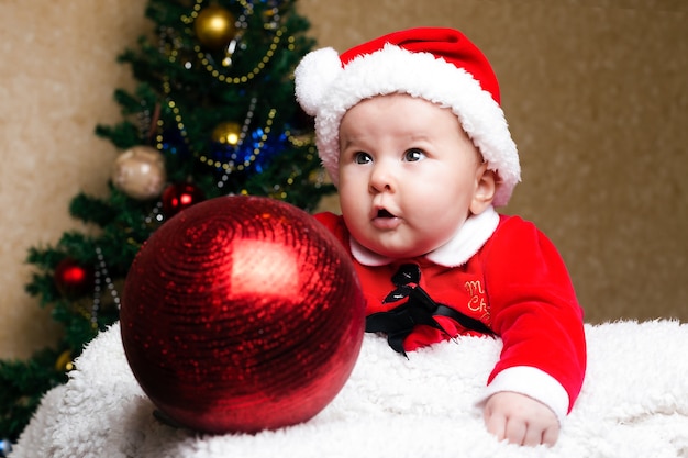 Lindo bebé navideño con traje de santa claus con una divertida mirada de sorpresa, árbol de navidad en la pared