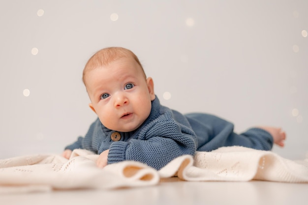 Lindo bebé con ojos azules en mono de punto azul yace boca abajo sobre fondo sólido a cuadros gris