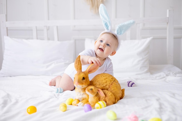 Lindo bebé con orejas de conejo y huevos coloridos en una cama blanca en casa jugando al pequeño bebé rubio con un conejo feliz concepto de pascua