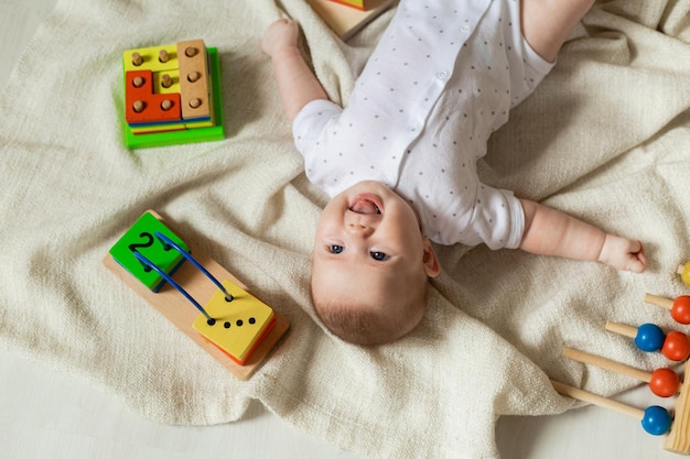 Un lindo bebé recién nacido con ropa ligera está jugando con juguetes educativos tirados en el suelo sobre una manta de lino gris. vista superior. productos para niños. feliz infancia y maternidad. espacio para texto