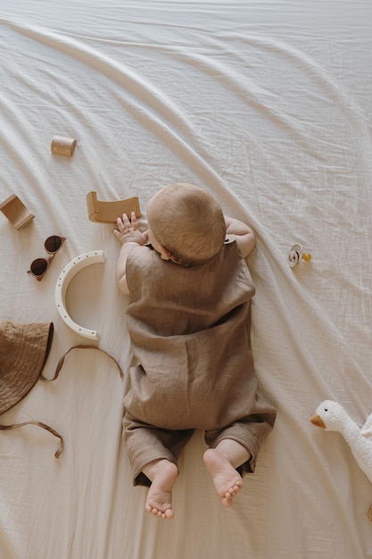 Lindo bebé recién nacido en traje de lino marrón acostado y jugando con juguetes en sábanas de cama beige Composición estética de moda para bebés Vista superior plana