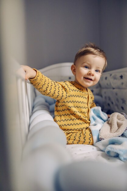 Lindo bebé rubio alegre sentado en su cuna por la mañana y está feliz de ver a sus padres. También está esperando su delicioso desayuno.