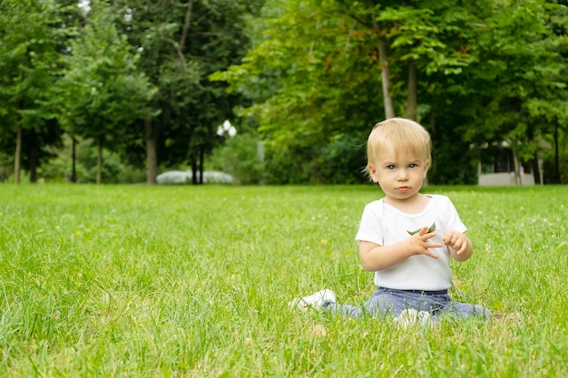 Lindo bebé rubio caucásico de aproximadamente 1 año de edad, sentado en la hierba en el parque, en verano, jugando con la hoja, riendo, mirando a la cámara. Niño pasar tiempo al aire libre en la naturaleza. Copiar espacio para texto.