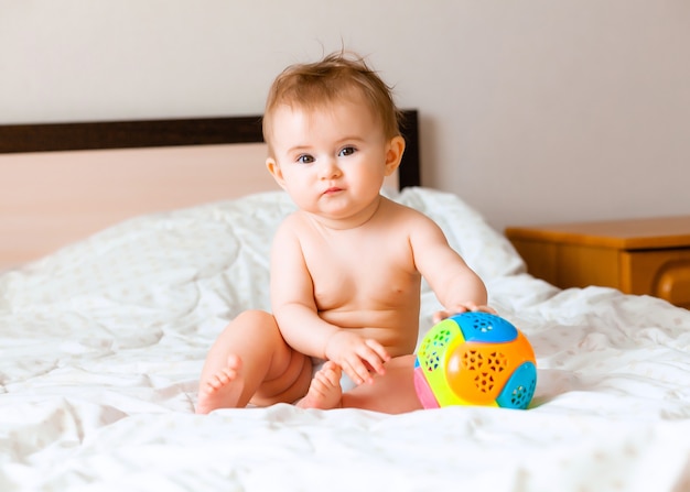 Lindo bebé rubio jugando con una pelota sentada en la cama en el dormitorio. Bebé feliz de 6 meses jugando con una pelota