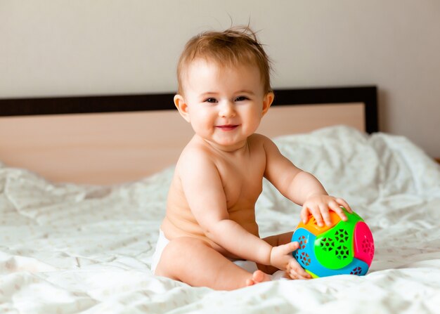 Lindo bebé rubio jugando con una pelota sentada en la cama en el dormitorio. Bebé feliz de 6 meses jugando con una pelota