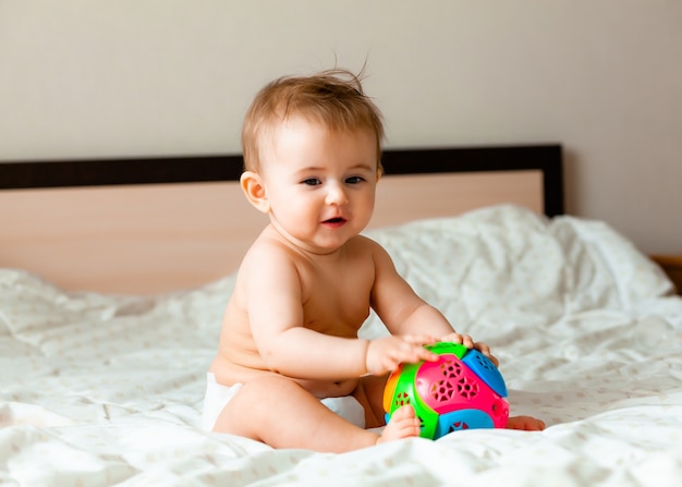 Lindo bebé rubio jugando con una pelota sentada en la cama en el dormitorio. Bebé feliz de 6 meses jugando con una pelota