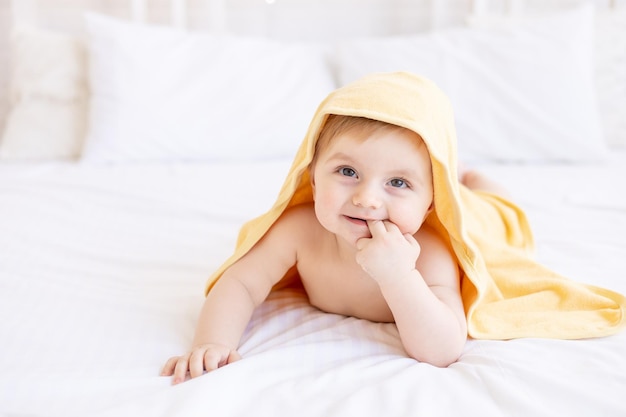 Lindo bebé rubio con una toalla amarilla en la cabeza después de un baño o bañándose en una cama blanca en casa en una habitación luminosa sonríe el concepto de cuidado infantil e higiene